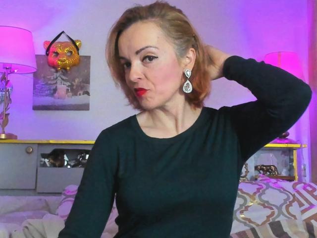 KateRouge with red lipstick, black top, and diamond earrings, hand in hair, purple and pink room, pink lamp, tiger mask, and bed with geometric pattern.