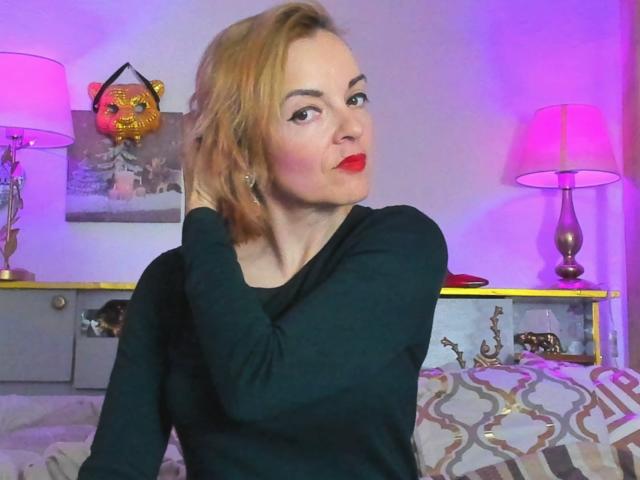 KateRouge with red lipstick, black top, and blonde hair, touching her head, sitting on a bed with pink and purple lamps, and a lion mask on the wall.