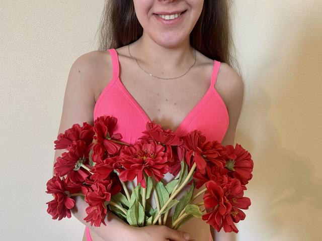 JoannaSia with red daisy flowers, pink bra, smiling, holding bouquet, long brown hair, subtle necklace, white wall, cute, medium tits, natural light.