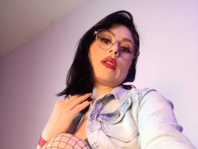 VioletaWhitee with black hair, glasses, red lipstick, denim jacket, and fishnet top, showing her tits, hand on collar, looking down at camera.