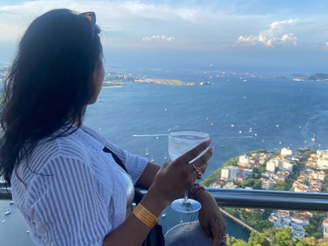 ElizabethMacy with dark hair, white striped shirt, holding a clear glass of water, looking at a coastal view, wearing yellow wristband, sitting by a glass railing.
