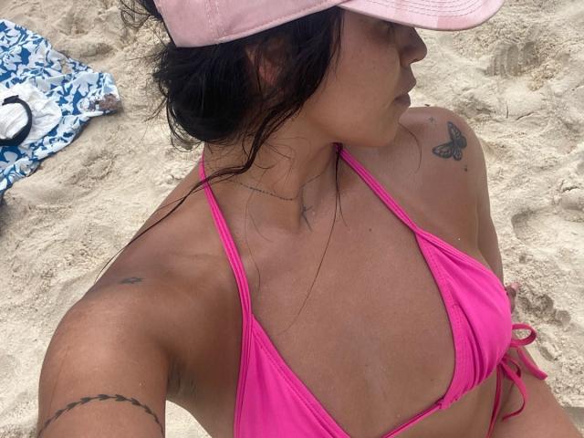 Elizabeth Macy, con piel morena, cabello negro bajo un gorro rosa, traje de baño rosa, mostrando pequeños senos, un tatuaje de mariposa en el hombro derecho y tatuajes en los brazos, sentada en una playa arenosa..