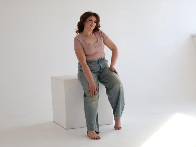 StaceyLex sits on a white cube, barefoot, wearing a pink top and blue jeans. She has curly brown hair, a cute smile, and small tits. Her jeans are slightly dirty.