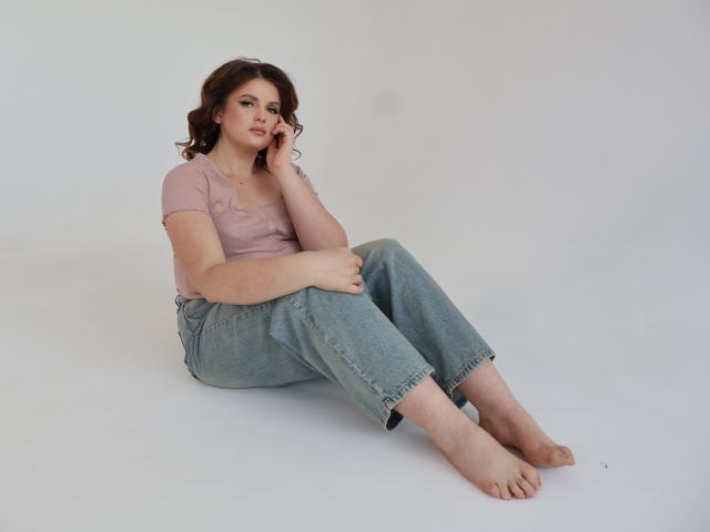 StaceyLex sits on the floor with legs crossed, wearing a pink top and blue jeans, barefoot, her brown curly hair, hand on cheek, looking slightly bored.