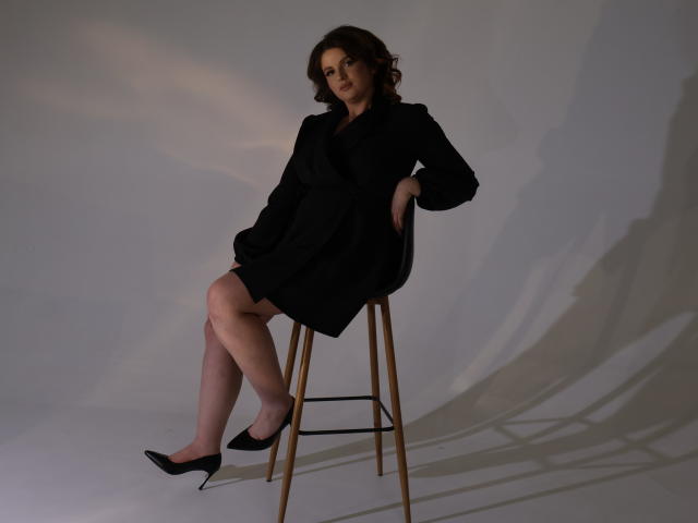 StaceyLex with curly brown hair, wearing a black dress and black heels, sitting on a stool with her ass slightly raised, hands in pockets, legs crossed.