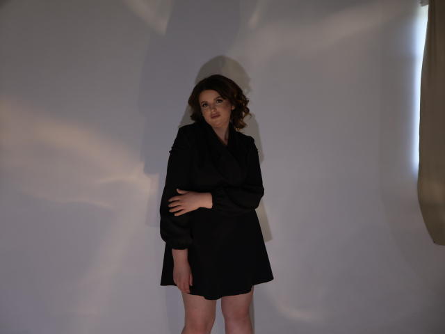 StaceyLex, com cabelos castanhos cacheados, vestindo um vestido preto, em pé com a mão direita sobre a barriga, sombra em uma parede branca, olhando ligeiramente para o lado..