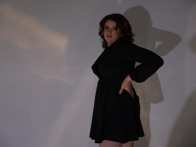 StaceyLex está de pie con una mano en la cadera, vestida con un vestido negro, con una sombra proyectada sobre una pared blanca, cabello castaño rizado, maquillaje sutil, expresión segura, iluminación suave..