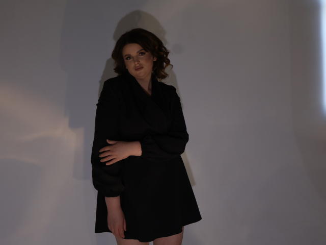 StaceyLex met golvend bruin haar, gekleed in een zwarte jurk, staat met één hand op haar buik, er is een schaduw op de muur, ze kijkt iets opzij, zachte belichting..