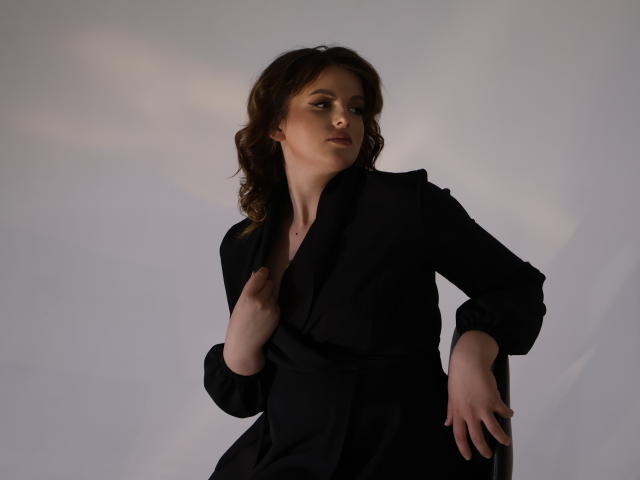 StaceyLex with wavy brown hair, wearing a black blazer, hand on chest, other hand on chair, looking slightly to the side, soft lighting, subtle shadow, confident pose.