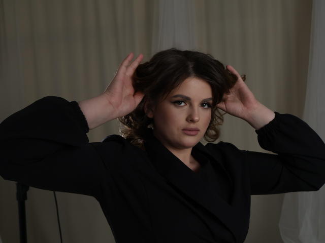 StaceyLex with curly brown hair, wearing a black shirt, hands in her hair, serious expression, standing in a dimly lit room. Her tits are small and she's looking at the camera.