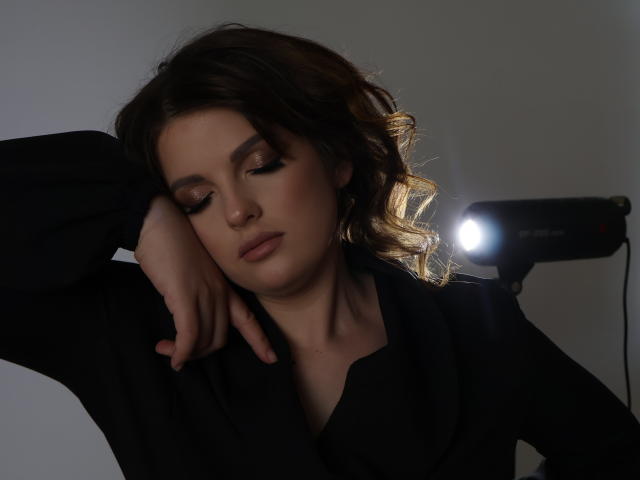 StaceyLex, les yeux fermés, cheveux noirs et ondulés, chemise noire, bras reposant sur la tête, lumière douce provenant d'un flash sur la droite, l'air détendue et légèrement séduisante..