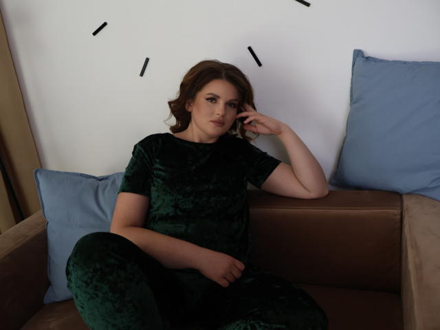 StaceyLex, con cabello castaño ondulado, vestida con un vestido de terciopelo verde, sentada en un sofá marrón con cojines azules, con la mano en la cara, luciendo ligeramente molesta..