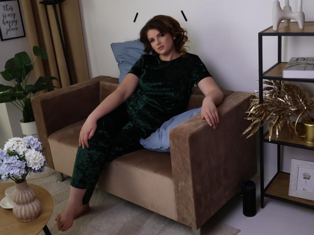 StaceyLex with curly brown hair, wearing green velvet pants and matching top, sitting on a brown suede sofa, arms relaxed, legs crossed, blue pillow behind her.