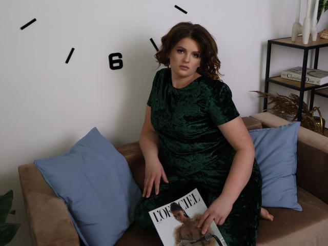 StaceyLex with curly brown hair, wearing a green velvet dress, sits on a beige sofa, holding a magazine with a photo of a man, between blue pillows.