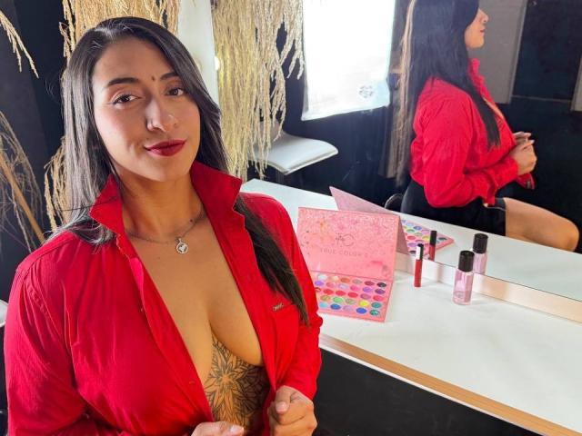 ZaraBeicker with long dark hair, red shirt, deep cleavage, showing her tits and tattoo, sitting at makeup table, reflection in mirror, pink perfume, stickers, and brushes.