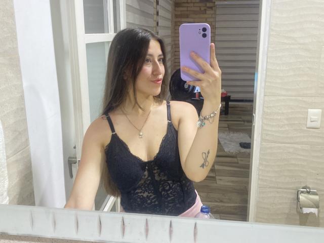 KatiaRogers with long dark hair, wearing a black lace bra, holding a purple phone, mirror selfie, visible tattoo on right arm, pink pants, smiling, white wall background.