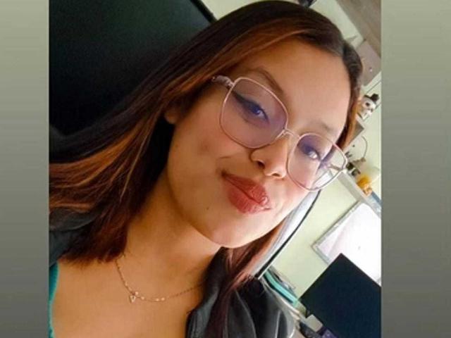 Female photo of MalejaI with brown hair, glasses, light skin, and red lipstick, making a pouty face, wearing a green top and a gold necklace.