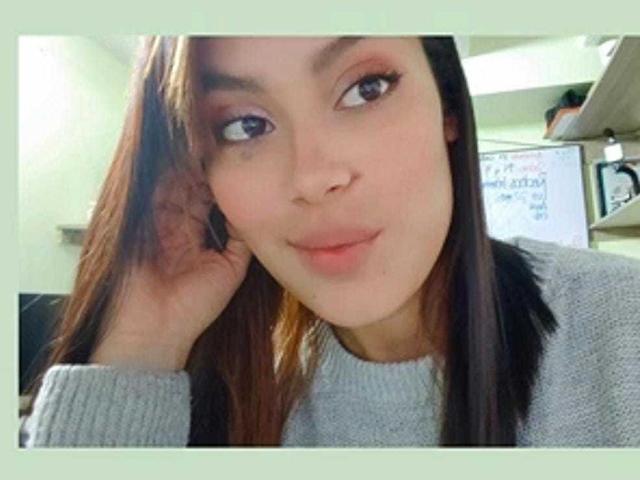 MalejaI has a cute face, light brown skin, and long brown hair, wearing a gray sweater, with a subtle smile, and a small mole under her right eye.