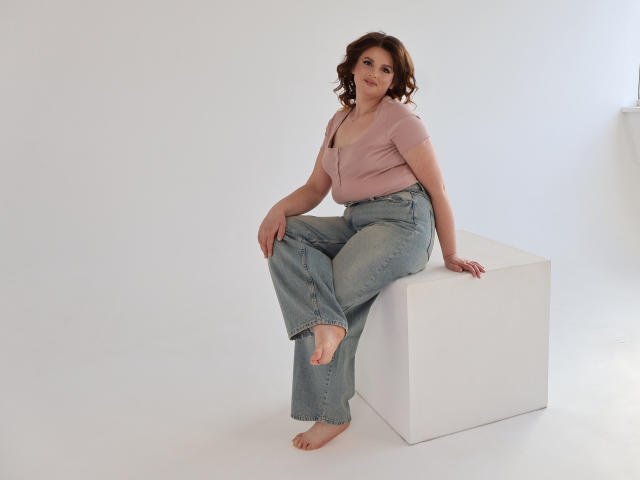 StaceyLex with curly brown hair, pink top, and blue jeans, sitting barefoot on a white cube, smiling at the camera, showing her bare toes and relaxed pose.