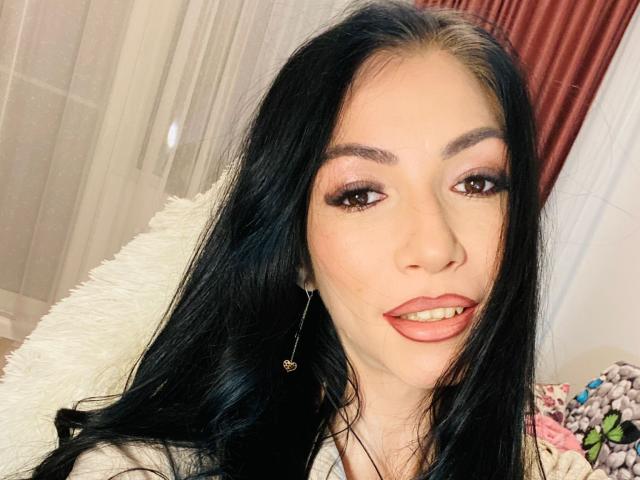Crazysin has long black hair, brown eyes, pink eyeshadow, red lipstick, and is wearing a white fluffy chair. Her tits are small and her ass is not visible.
