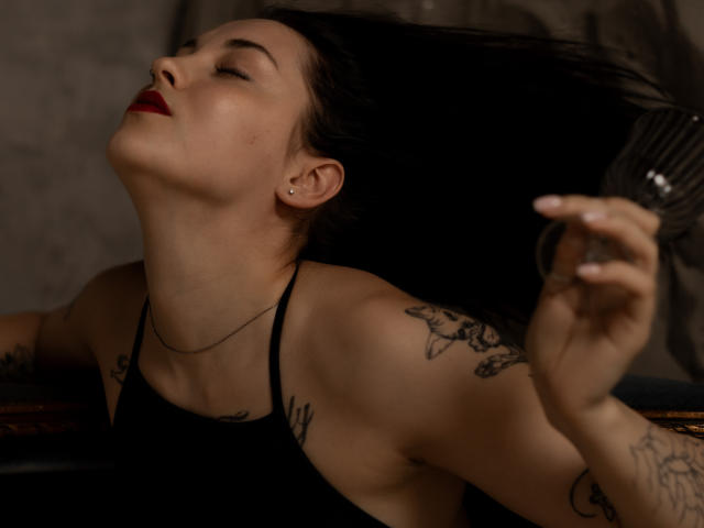 AngelinaSwt's arm tattooed with floral designs, raised, holding a metal brush, her bare shoulder and upper back visible, dark fabric partially covering her body.