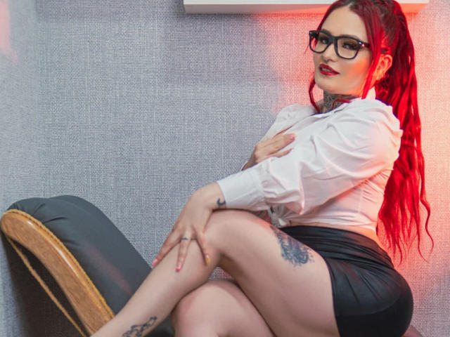 Isabella Dankworth, con i capelli rossi, gli occhiali, la camicia bianca, la gonna nera, seduta su una sedia, con le gambe incrociate, una mano sul petto, un tatuaggio sulla coscia, illuminazione rossa, che guarda la fotocamera, sexy..