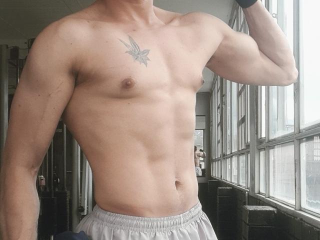 FlacoVergon69 is a muscular, shirtless guy with a bird tattoo on his chest, small tits, and white pants, standing by a window, showing his defined abs and arm.