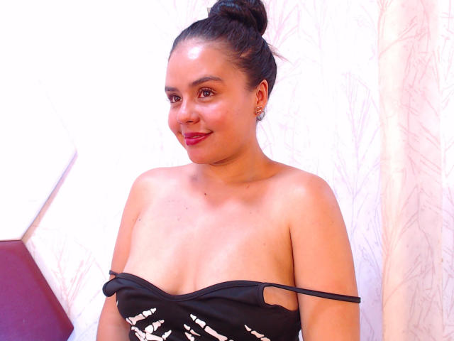 ArielleWilde with dark hair in a bun, red lipstick, and black strapless dress showing her small tits, standing against a white wall, smiling slightly.