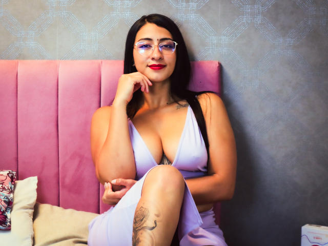 ZaraBeicker with big tits, red lipstick, glasses, and black hair, wearing a low-cut white dress, sitting on a pink bed, touching her chin, showing a tattoo on her thigh.