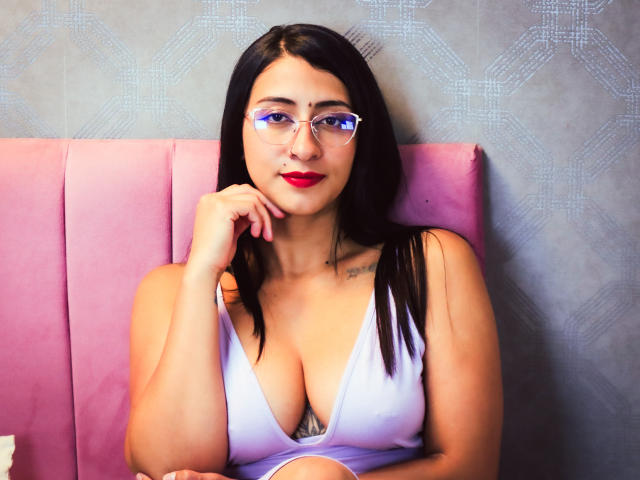 ZaraBeicker with black hair, red lipstick, glasses, and white low-cut top showing big tits, resting chin on hand, pink cushioned seat, tattoo on right shoulder.