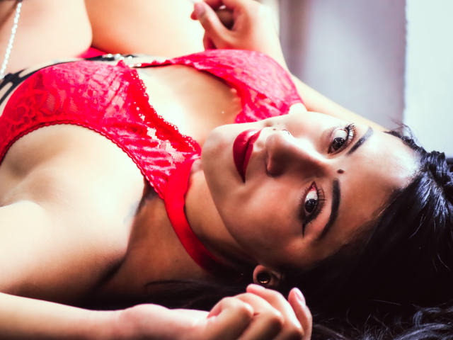 ZaraBeicker with red lace lingerie, lying down, looking up with red lipstick, black hair, and a cute mole on her right cheek, showing her tits and a sexy smile.