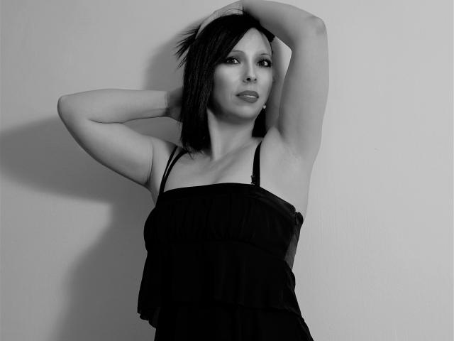 LauraSin with arms raised, black dress, small tits, short dark hair, light skin, serious expression, black and white photo, standing against plain background.