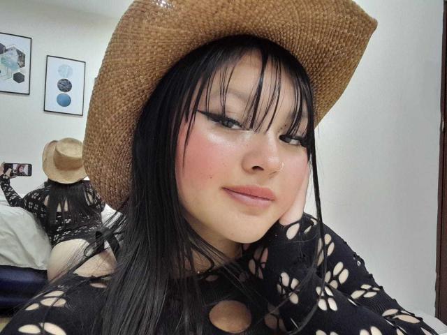 MiaRoots with black hair, wearing a woven hat, black lace top, and pink blush, winking, nose pierced, touching cheek, bed and wall art in background.