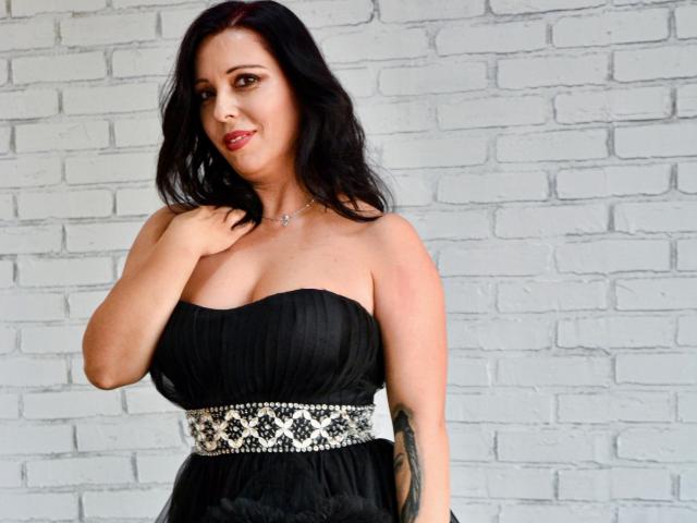LauraSin with dark wavy hair, red lipstick, black strapless dress, white patterned belt, tattoo on right arm, standing against white brick wall, hand on chest, smiling.