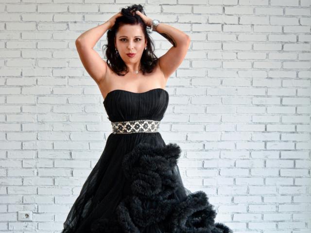 LauraSin with hands in dark hair, wearing a strapless black dress with a fur skirt, silver belt, and bracelet, standing against a white brick wall.