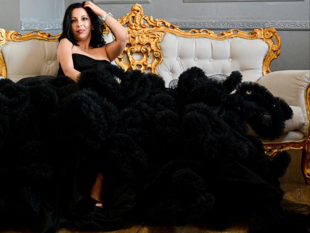 LauraSin with black fur blanket, sitting on ornate gold and white sofa, wearing strapless black dress, hand in hair, looking at camera, her tits partially covered by fur.