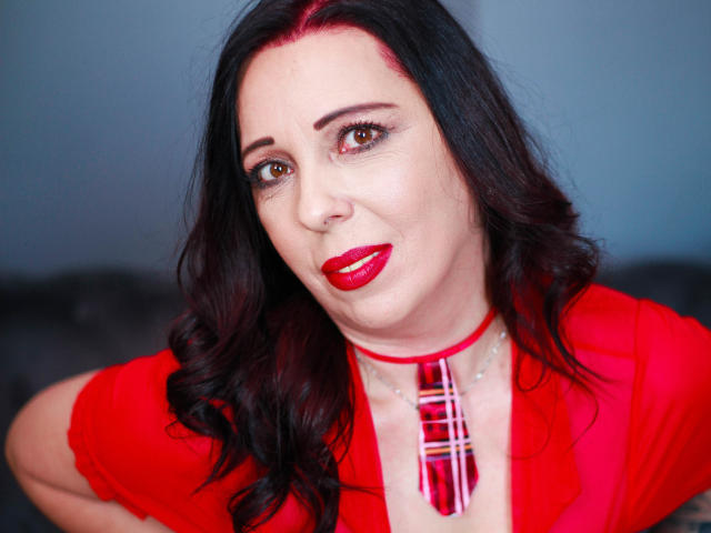 LauraSin with red lipstick, dark wavy hair, red dress, and red plaid necklace, looking at camera, medium shot, red highlights in hair, blue background.