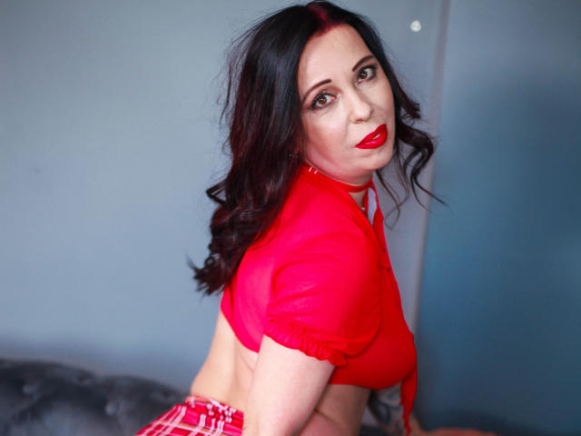 LauraSin with red lipstick, dark wavy hair, red crop top, red plaid skirt, looking back, showing her tits, red ribbon in her hair, blue-gray wall in background.