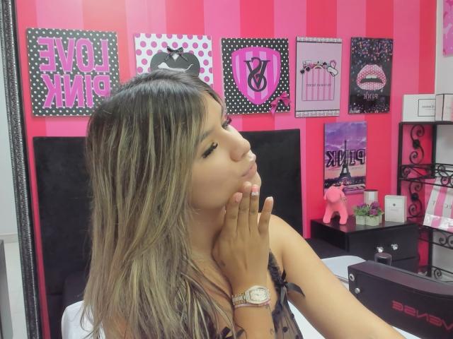 SashaPolet with long, blonde-highlighted hair, pouting lips, and hand near face, wearing a black top with a ribbon, in a pink and black room, with a watch on wrist.