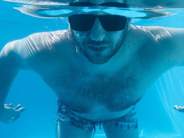 Underwater shot of AxelCroftAndFordFairlane, a hairy-chested dude with sunglasses, leaning forward, wearing checkered swim trunks, showing a slightly open mouth and blue-tinted skin.