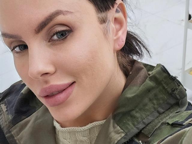 AxelCroftAndFordFairlane has a cute face, blue eyes, and is licking her pink lips. She's wearing a green camo jacket with a beige sweater underneath.