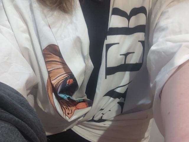Ajiki69's shirt with a colorful graphic, black text, and slightly loose fit, covering their tits. Visible gray fabric on the left, Ajiki69's arm on the right.