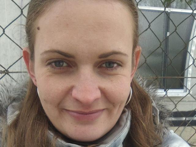 Ajiki69's face with a small mole on the left side, gray eyes, and a subtle smile. She has brown hair in a braid, wearing a fur-trimmed jacket and silver hoop earrings.