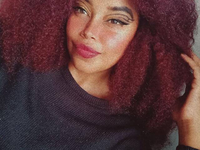 DalilaHeinz with curly red hair, dark eyebrows, winged eyeliner, pink lipstick, and a black sweater, touching her hair. Her ass is not visible.