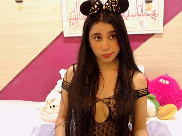 TiffannyRose, com longos cabelos pretos, vestindo um vestido preto com recortes e orelhas do Mickey, sentada em uma cama com brinquedos de pelúcia, tem um rosto fofo e seios pequenos..