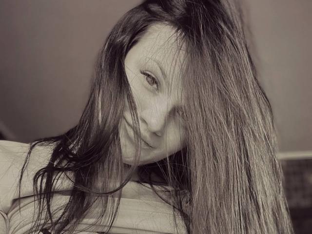 CamilleXMyr con lunghi capelli castani lisci che le coprono gli occhi, sorride leggermente, indossa una maglietta di colore chiaro, in una foto in tonalità seppia, il suo viso è parzialmente nascosto dai capelli..