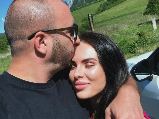 AxelCroftAndFordFairlane with bald head and black glasses kissing forehead of FordFairlane with shiny skin, dark straight hair, and pink lips, standing next to a white car.