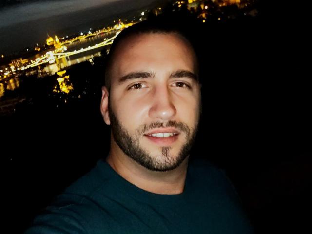 AxelCroftAndFordFairlane has a shaved head, beard, and slight mustache, wearing a dark green shirt, smiling at the camera with a dimly lit cityscape in the background.