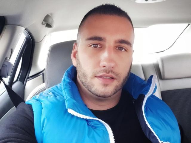 AxelCroftAndFordFairlane, with short buzzed hair and stubble, wearing a blue puffy vest over a black shirt, sitting in a car. His face is slightly sweaty.