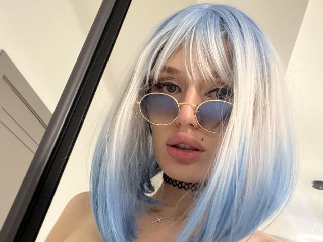 SweetMirakle with blue and white hair, gold aviator sunglasses, black choker, and pink lips, showing a small beauty mark on her left cheek, with a cross necklace.